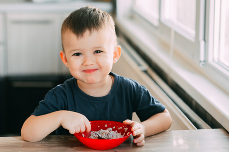 A child in the kitchen eating their own oatmeal with a red plateの写真素材