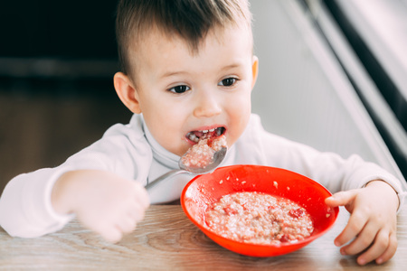 A child in the kitchen eating their own oatmeal with a red plate