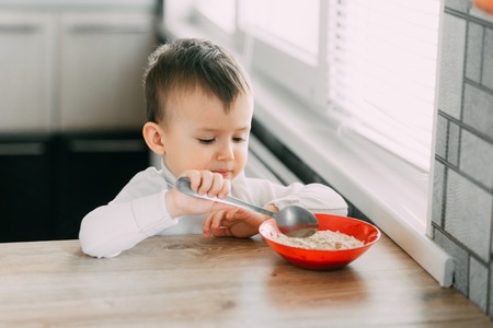 A child in the kitchen eating their own oatmeal with a red plateの写真素材