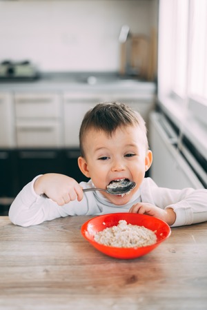 A child in the kitchen eating their own oatmeal with a red plateの写真素材