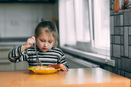 girl eating pasta with sausage in the kitchen in a striped jacketの写真素材
