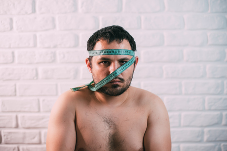 Portrait of bearded frustrated fat man with green measuring tape around his mouth, healthy eating conceptの写真素材