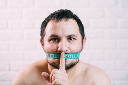 Portrait of bearded frustrated fat man with green measuring tape around his mouth, healthy eating conceptの写真素材