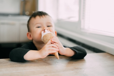 A child in a dark-blue t-shirt in the bright kitchen eating a waffle ice cream cone in the summer houseの写真素材