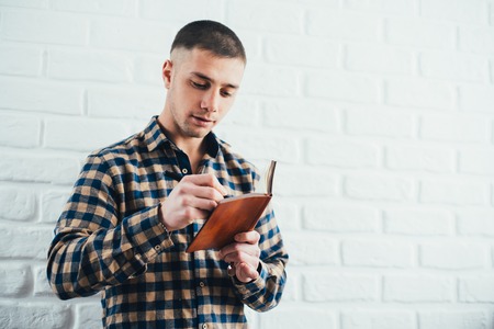 Businessman or student in a plaid shirt against a brick white wall writes in a notebookの写真素材