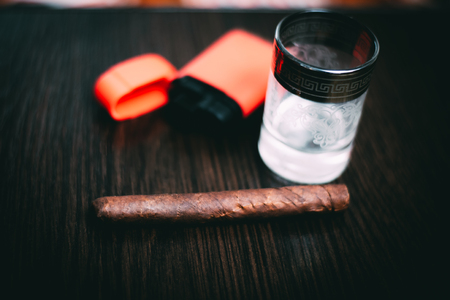 On a dark wooden background is a glass of vodka next to a cigar and a lighterの写真素材