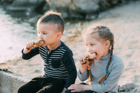 Children, brother and sister have fun eating chicken Shin on the beach near the sea and rocks, very appetizingの写真素材