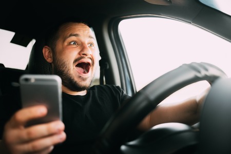 Young man sitting in his car screaming distracted by smartphone textingの写真素材