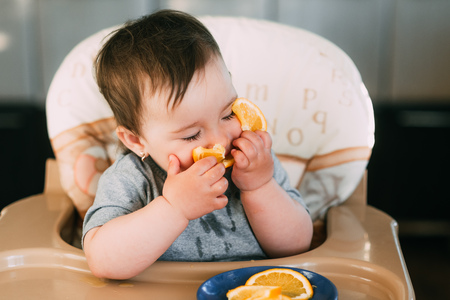 a little girl in a blue t-shirt and a blue plate sitting in a child's chair eating an orangeの写真素材