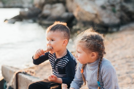 Children, brother and sister have fun eating chicken Shin on the beach near the sea and rocks, very appetizingの写真素材