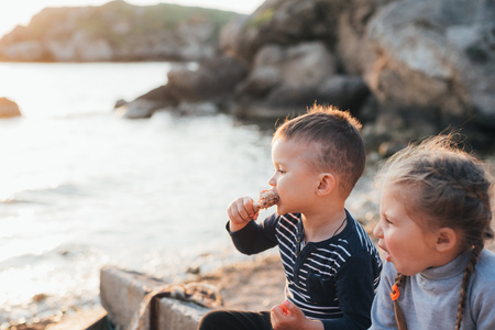 Children, brother and sister have fun eating chicken Shin on the beach near the sea and rocks, very appetizingの写真素材