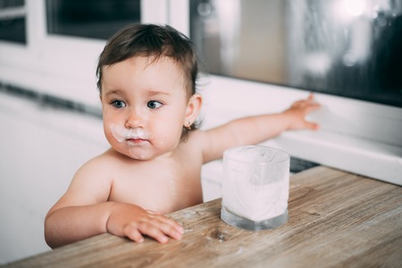 A little girl in the kitchen drinking milk or yogurt from a glass is very appetizing and greedy all face dirty in milkの写真素材