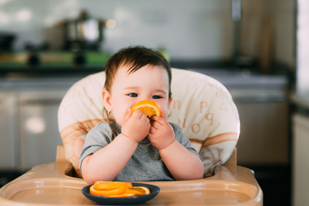 a little girl in a blue t-shirt and a blue plate sitting in a child's chair eating an orangeの写真素材