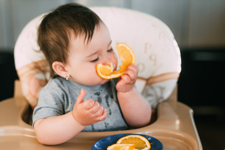 a little girl in a blue t-shirt and a blue plate sitting in a child's chair eating an orange
