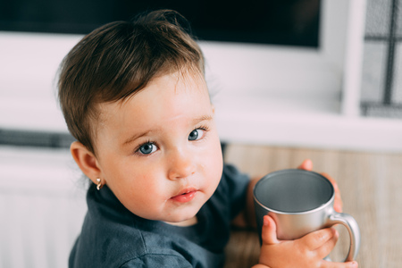 A little girl in the kitchen drinking water from a silver mug very greedily, thirsty