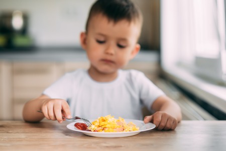 child day in the kitchen eating an omelet with sausage and ketchup in a white t-shirtの写真素材