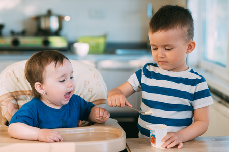 Kids in the kitchen, brother feeds baby sister yogurt with a spoonの写真素材