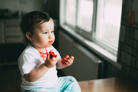 A little girl child in a white t-shirt climbed on the table sitting and eating strawberriesの写真素材