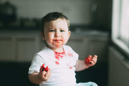 A little girl child in a white t-shirt climbed on the table sitting and eating strawberriesの写真素材