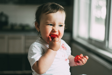 A little girl child in a white t-shirt climbed on the table sitting and eating strawberriesの写真素材