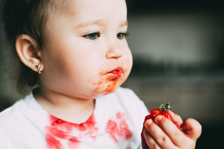 A little girl child in a white t-shirt climbed on the table sitting and eating strawberriesの写真素材