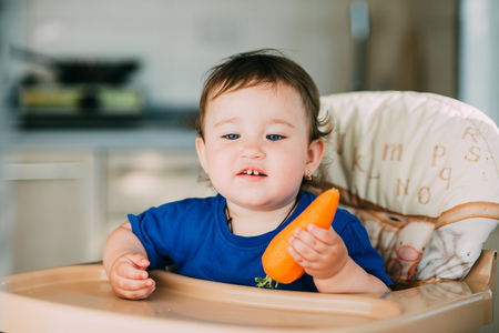 A little funny girl, a child in a high chair eating a carrot in the afternoon in the kitchenの写真素材