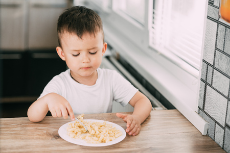 a child in the kitchen during the day eating pasta in a spiral in a white t-shirt at the tableの写真素材