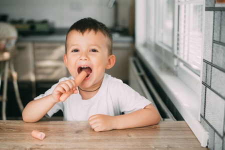 Portrait of a adorable little boy in a white t-shirt at the table eating a sausageの写真素材