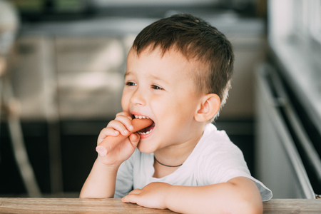 Portrait of a adorable little boy in a white t-shirt at the table eating a sausageの写真素材
