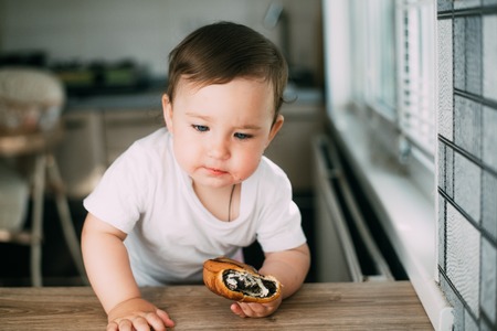 a little girl in the kitchen during the day eating a bun with poppy seeds is very appetizing and...