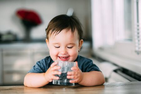 Cute little girl sitting in a baby chair in the kitchen and drinking waterの写真素材