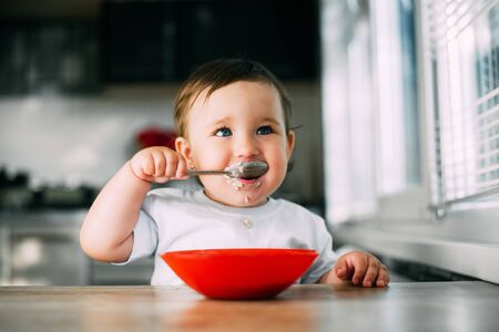 Funny little girl in the kitchen eating oatmeal porridge from a red plate all dirtyの写真素材
