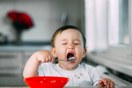Funny little girl in the kitchen eating oatmeal porridge from a red plate all dirtyの写真素材