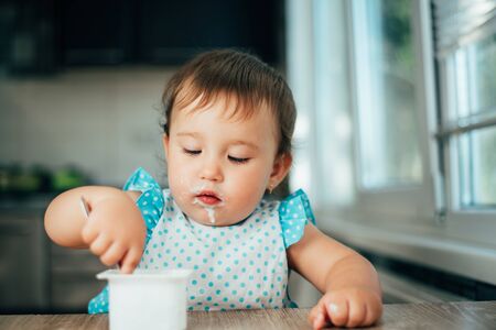 Cute, funny girl eating yogurt during the day in the kitchen in a blue dressの写真素材
