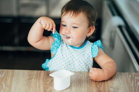Cute, funny girl eating yogurt during the day in the kitchen in a blue dressの写真素材