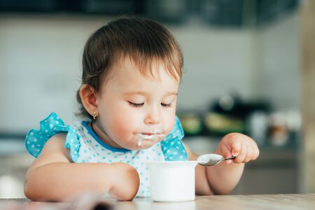 Cute, funny girl eating yogurt during the day in the kitchen in a blue dressの写真素材