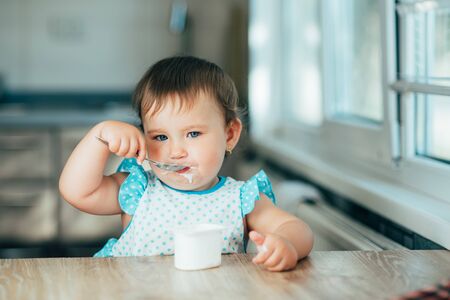 Cute, funny girl eating yogurt during the day in the kitchen in a blue dressの写真素材