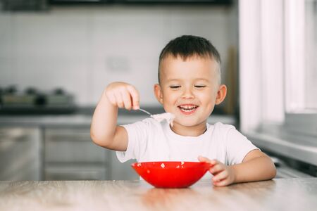 Little boy in the kitchen eating oatmeal from a red plateの写真素材