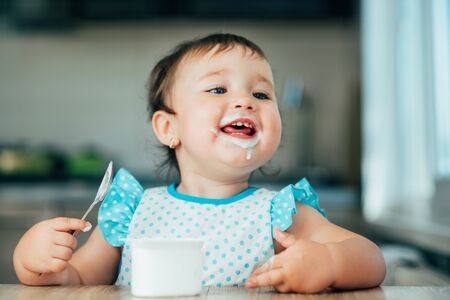 Cute, funny girl eating yogurt during the day in the kitchen in a blue dressの写真素材