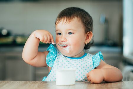Cute, funny girl eating yogurt during the day in the kitchen in a blue dressの写真素材