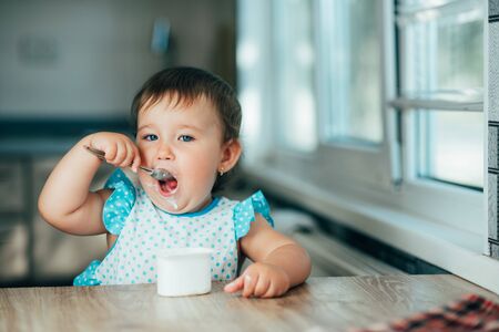 Cute, funny girl eating yogurt during the day in the kitchen in a blue dressの写真素材