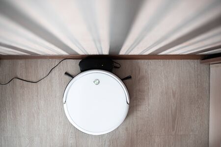 white robot vacuum cleaner in the interior on a light laminate is charging near the wallの写真素材
