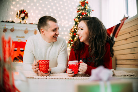 Cute couple on background of Christmas tree and fireplace lying on stomach with red mugsの写真素材