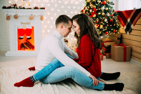 Cute couple on the background of Christmas tree and fireplace sitting on the floor hugging very close and lovingの写真素材