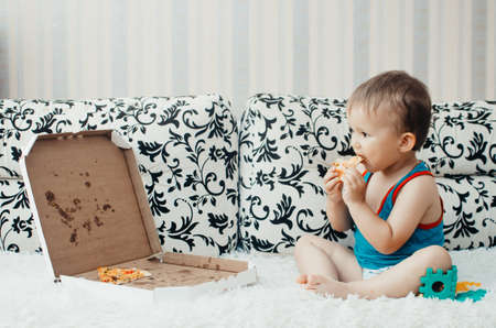 The boy eats a piece of delicious pizza sitting on the couch, shirtless, with a naked torsoの写真素材