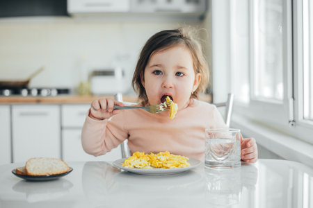 Baby in the afternoon in the white light kitchen in a pink sweater eating an omeletの写真素材