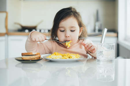 Baby in the afternoon in the white light kitchen in a pink sweater eating an omeletの写真素材