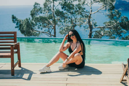 a girl in black sitting by the pool on a beautiful background of the sea and mountains, with braided...