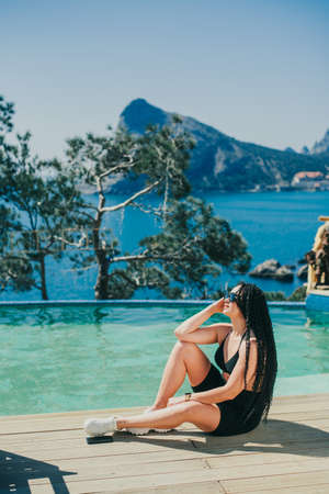 a girl in black sitting by the pool on a beautiful background of the sea and mountains, with braided...