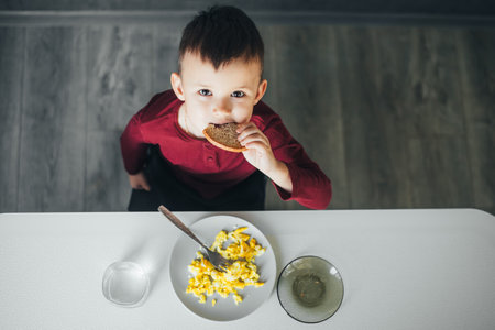 A beautiful little boy in the kitchen is eating a piece of bread, and an omeletの写真素材